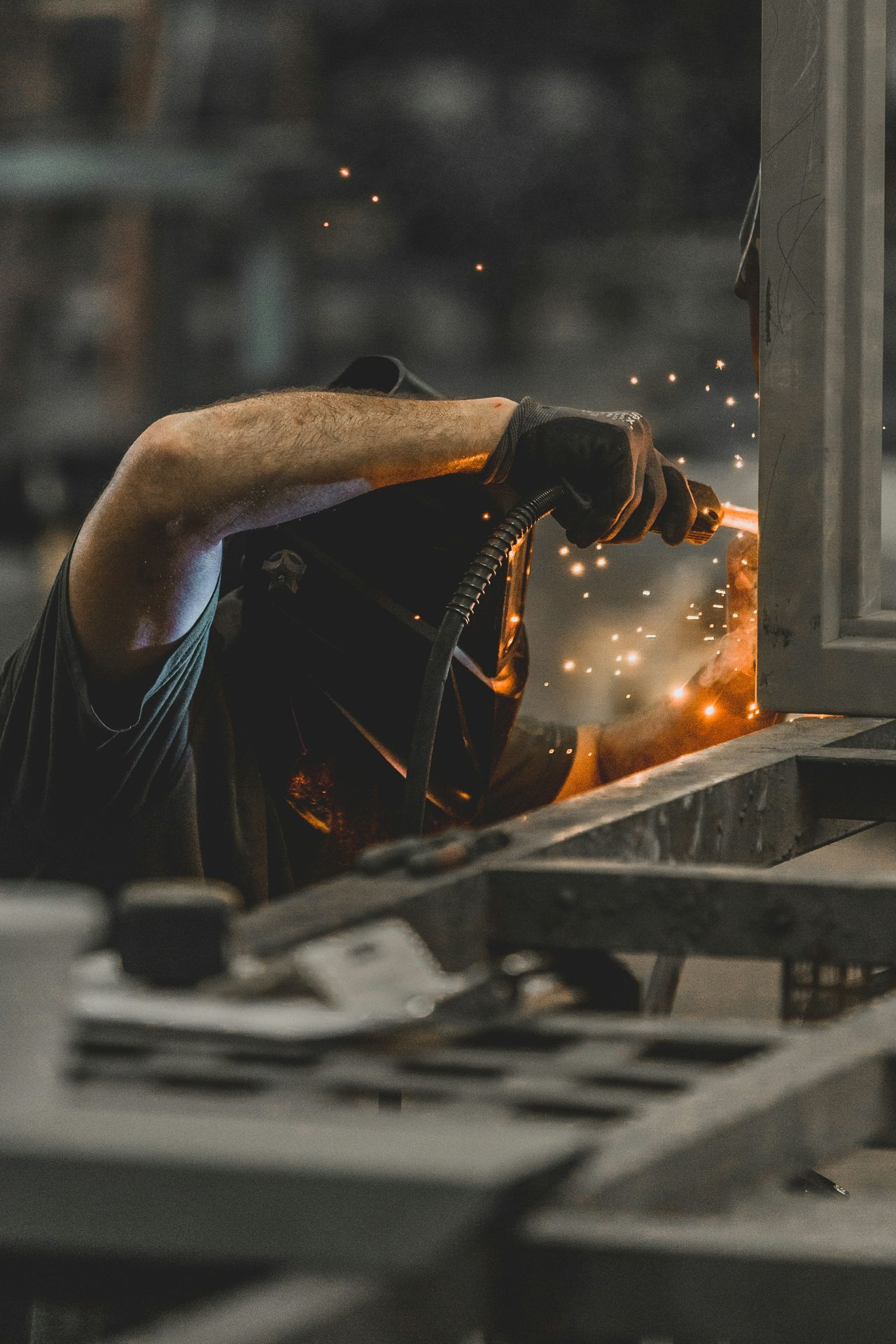 Un soudeur est penché sur une structure métallique, dans un atelier industriel. On le voit de profil, le visage caché derrière un masque de soudure sombre. Il porte un t-shirt sans manches gris foncé et un gant noir. Sa main tient une torche de soudage reliée à une gaine annelée. Au point de contact, une gerbe d'étincelles orange jaillit et se disperse dans l'air. La lumière chaude de la soudure éclaire son bras et le bord du masque. Au premier plan, des pièces de métal brut sont floues. L'arrière-plan, gris et sombre, laisse deviner l'atelier sans le montrer. Le contraste entre les tons froids du métal et l'éclat orangé des étincelles donne à l'image une ambiance à la fois brute et précise. Le format est vertical.