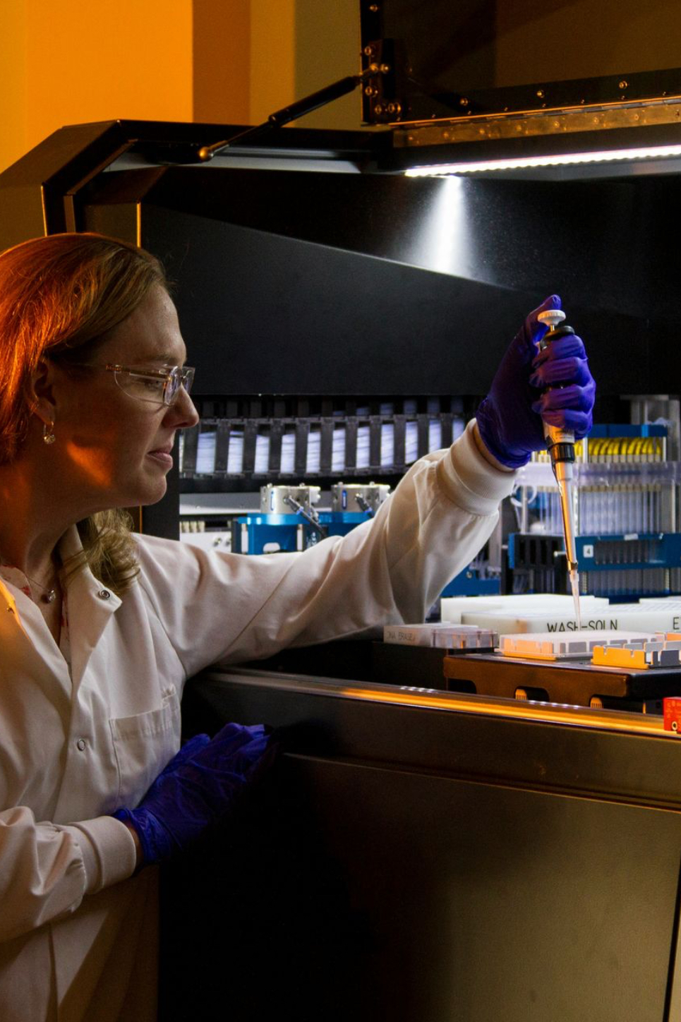 Une chercheuse travaille dans un laboratoire, penchée vers un grand automate de pipetage ouvert. On la voit de trois quarts. Elle porte une blouse blanche avec un prénom brodé sur la poitrine, des lunettes de protection transparentes et des gants en nitrile violets. D'une main, elle tient une micropipette au-dessus de plaques et de réservoirs blancs, dont certains portent des étiquettes techniques. De l'autre, elle s'appuie sur le rebord de la machine. Son regard est concentré sur le geste. L'intérieur de l'automate est éclairé par une rampe de lumière blanche qui révèle des rangées de cônes, des supports bleus et des mécanismes de précision. Le reste de la pièce baigne dans une lumière orange chaude, qui éclaire ses cheveux et sa blouse. Le contraste entre cette chaleur et l'éclairage froid de la machine met en valeur la rencontre entre le geste humain et la technologie de pointe. Le format est vertical