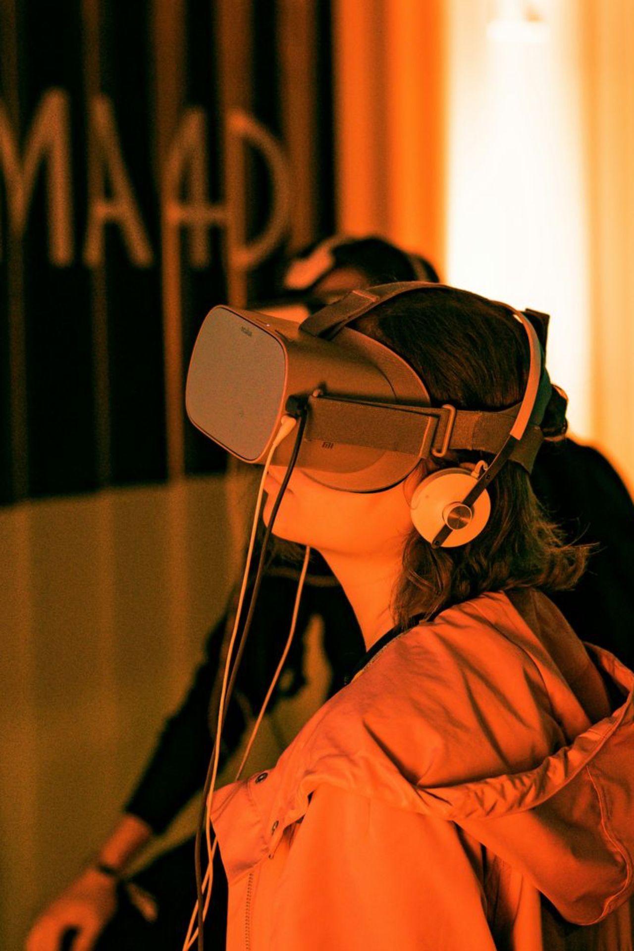 Une jeune femme vit une expérience de réalité virtuelle, debout dans une salle plongée dans une lumière orange. On la voit de profil, la tête légèrement levée. Elle porte un casque de réalité virtuelle gris sur les yeux et un casque audio blanc sur les oreilles. Des câbles fins descendent le long de son cou. Elle est vêtue d'une veste à capuche dont le tissu capte la lumière chaude. Derrière elle, une seconde personne, plus floue, porte elle aussi un casque. À gauche, un mur sombre affiche de grandes lettres blanches partiellement visibles. À droite, un écran diffuse un affichage pixelisé aux tons vert turquoise. Le contraste entre l'orange enveloppant et ce vert numérique renforce l'ambiance immersive et technologique de la scène. Le format est vertical.