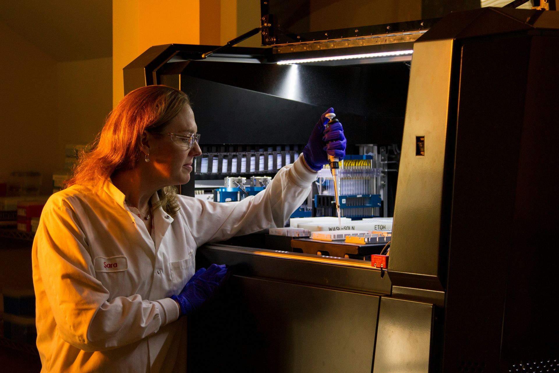 Une chercheuse travaille dans un laboratoire, penchée vers un grand automate de pipetage ouvert. On la voit de trois quarts. Elle porte une blouse blanche avec un prénom brodé sur la poitrine, des lunettes de protection transparentes et des gants en nitrile violets. D'une main, elle tient une micropipette au-dessus de plaques et de réservoirs blancs, dont certains portent des étiquettes techniques. De l'autre, elle s'appuie sur le rebord de la machine. Son regard est concentré sur le geste. L'intérieur de l'automate est éclairé par une rampe de lumière blanche qui révèle des rangées de cônes, des supports bleus et des mécanismes de précision. Le reste de la pièce baigne dans une lumière orange chaude, qui éclaire ses cheveux et sa blouse. Le contraste entre cette chaleur et l'éclairage froid de la machine met en valeur la rencontre entre le geste humain et la technologie de pointe. Le format est vertical