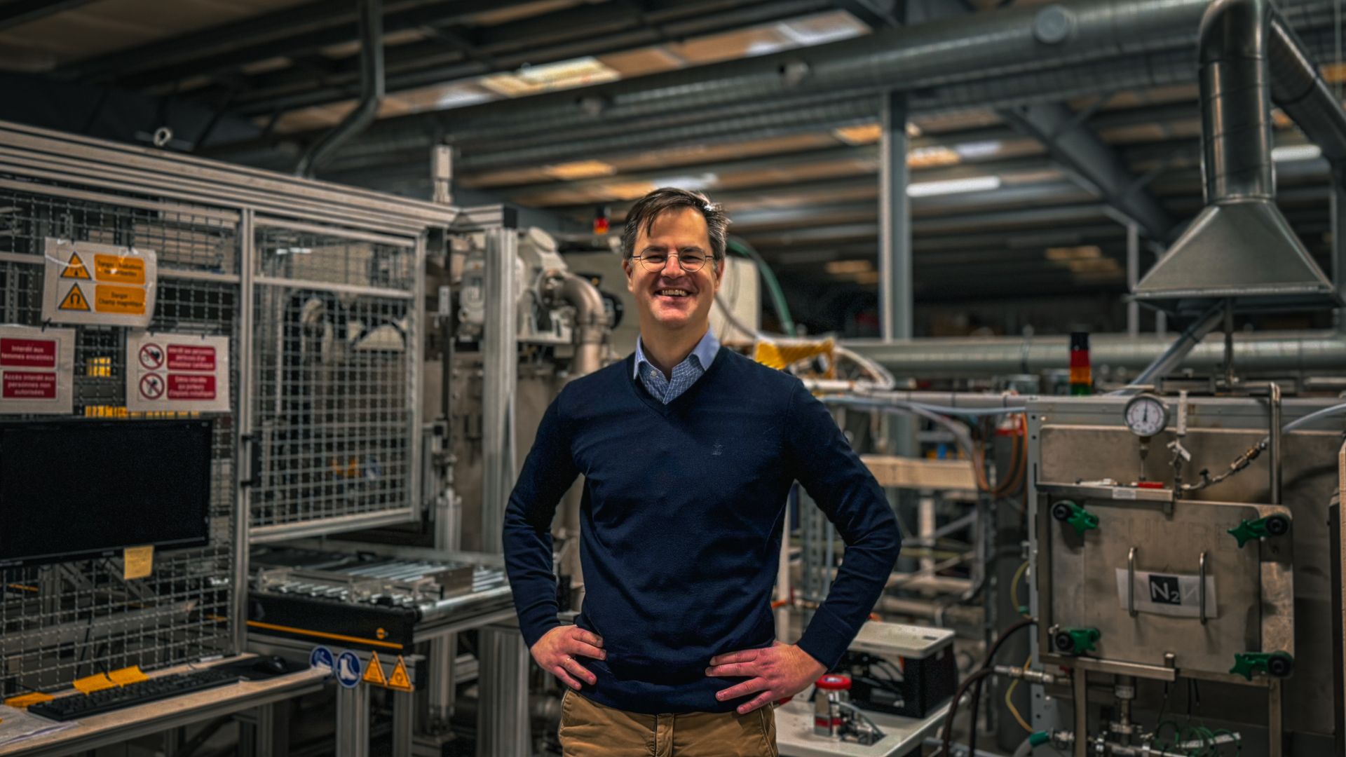 Antoine de Ryckel Antoine de Ryckel, CEO de Calyos, pose debout au milieu d'un atelier industriel. Il fait face à l'objectif, les mains sur les hanches, avec un large sourire. Il porte des lunettes rondes à fine monture, un pull bleu marine à col en V sur une chemise bleu clair, et un pantalon beige. Autour de lui, l'espace est dense et technique. À gauche, une machine protégée par des grilles métalliques affiche plusieurs panneaux de sécurité jaunes et rouges, à côté d'un écran et d'un clavier. À droite, une installation en inox équipée d'un manomètre et de vannes vertes porte une étiquette indiquant de l'azote. Au plafond, de larges gaines de ventilation et des structures métalliques traversent le cadre. Les tons froids et métalliques de l'atelier contrastent avec l'attitude détendue et assurée du dirigeant. L'image dégage une impression de confiance et d'ancrage dans l'outil industriel. Le format est horizontal.